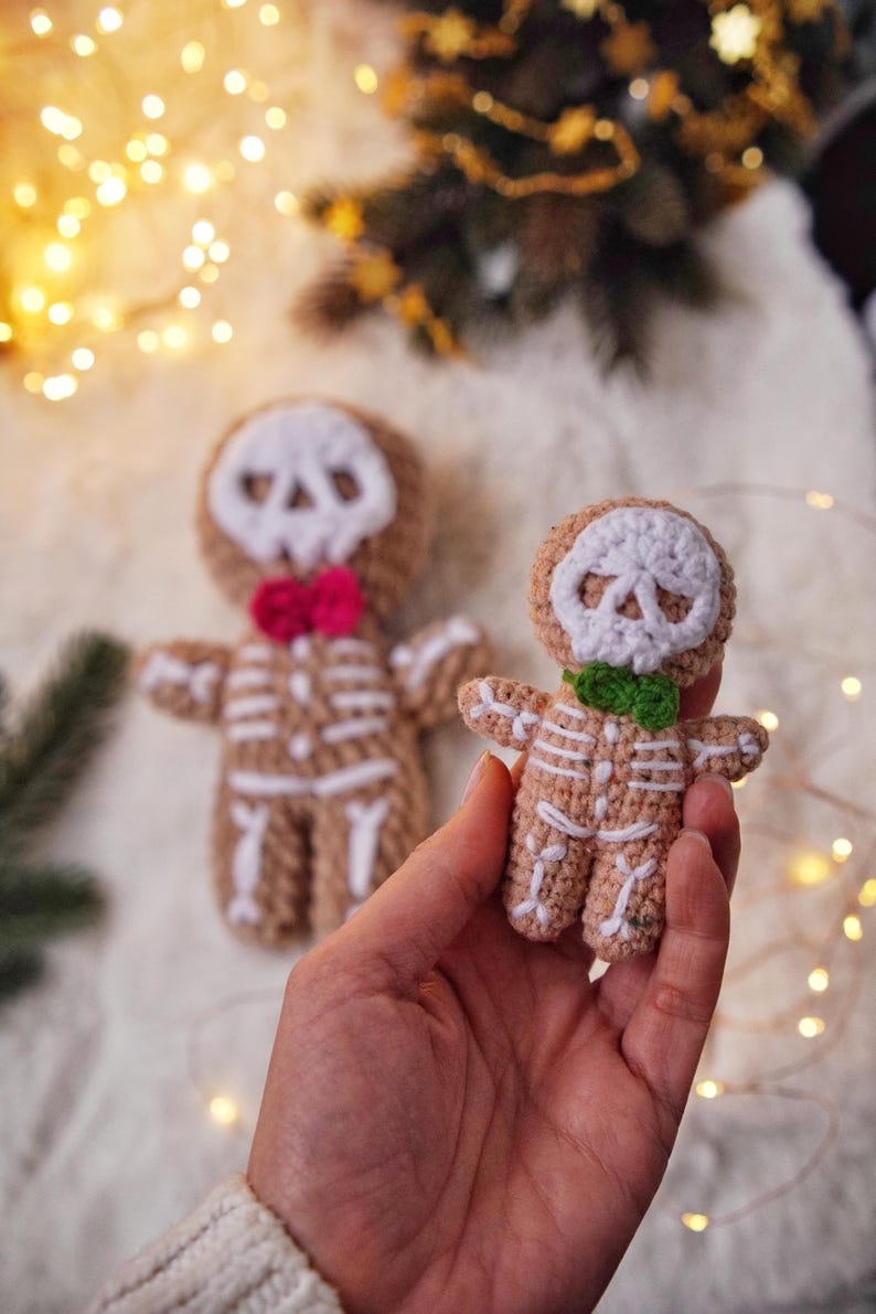 May include: Crocheted skeleton gingerbread men. The figures are tan with white skeleton detailing and skull faces. One has a pink bow tie, the other a green bow tie. The background includes a Christmas tree and fairy lights.