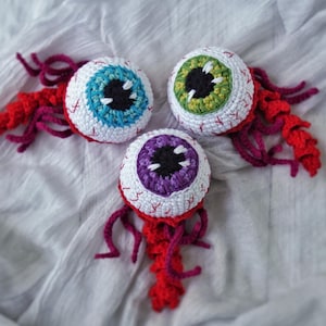 Puede incluir: Tres globos oculares de ganchillo hechos a mano con iris azules, verdes y morados. Cada ojo tiene una esclerótica blanca con costuras rojas, una pupila negra y zarcillos rojos y morados. Los ojos están sobre una tela blanca.
