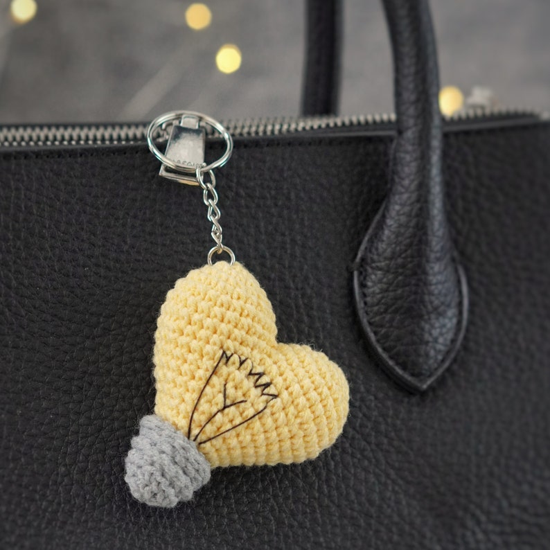 May include: A yellow crocheted heart-shaped lightbulb keychain with a gray crocheted base attached to a silver key ring. The keychain is hanging from a black leather handbag.