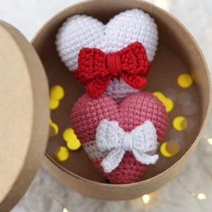 May include: Two crocheted hearts in a brown box. The top heart is white with a red bow. The bottom heart is pink with a white bow.
