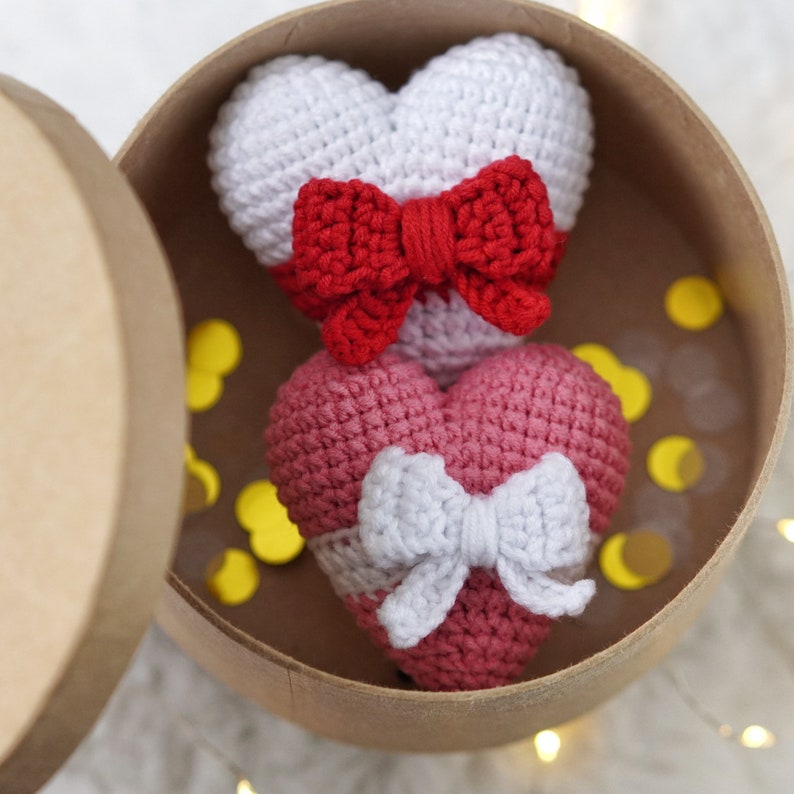 May include: Two crocheted hearts in a brown box. The top heart is white with a red bow. The bottom heart is pink with a white bow.
