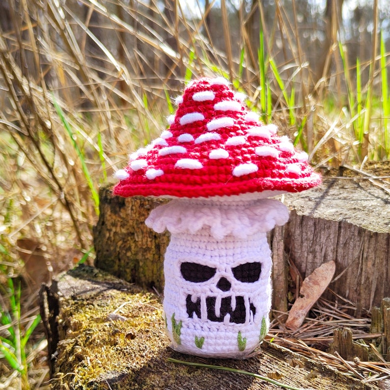 May include: A handmade crochet mushroom with a red cap dotted with white spots. The mushroom has a white stem with a skull face design and green accents. The mushroom is sitting on a mossy log in a natural outdoor setting.