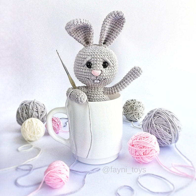 May include: A gray crocheted bunny rabbit sitting in a white ceramic mug. The bunny has black eyes, a pink nose, and a toothy smile. There are balls of yarn in various colors scattered around the mug.
