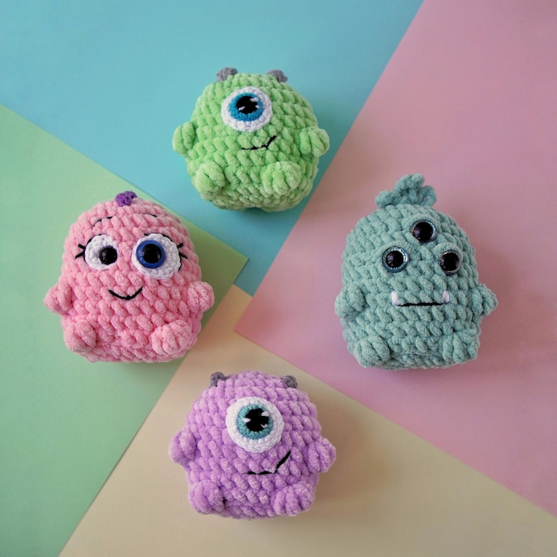 May include: Four crocheted plush toys shaped like monsters with different colors and eye designs. The monsters are green, pink, purple, and light green.