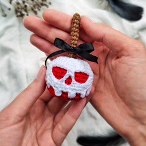 May include: A crocheted apple with a white skull design and a black ribbon. The apple is red and white and is held in a person's hand.