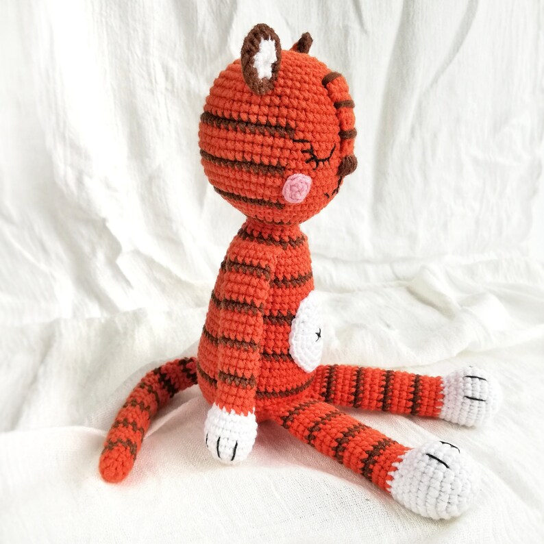 May include: A crocheted orange and brown striped cat toy with white paws and a pink nose. The cat is sitting with its tail curled behind it.