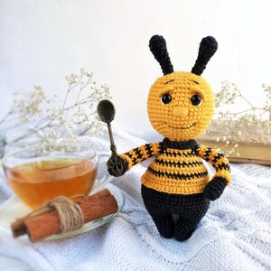 May include: A crocheted bee toy with black and yellow stripes, holding a small spoon. The bee has a smiling face with black eyes and a black nose. The bee is sitting on a white surface with a cup of tea and cinnamon sticks.