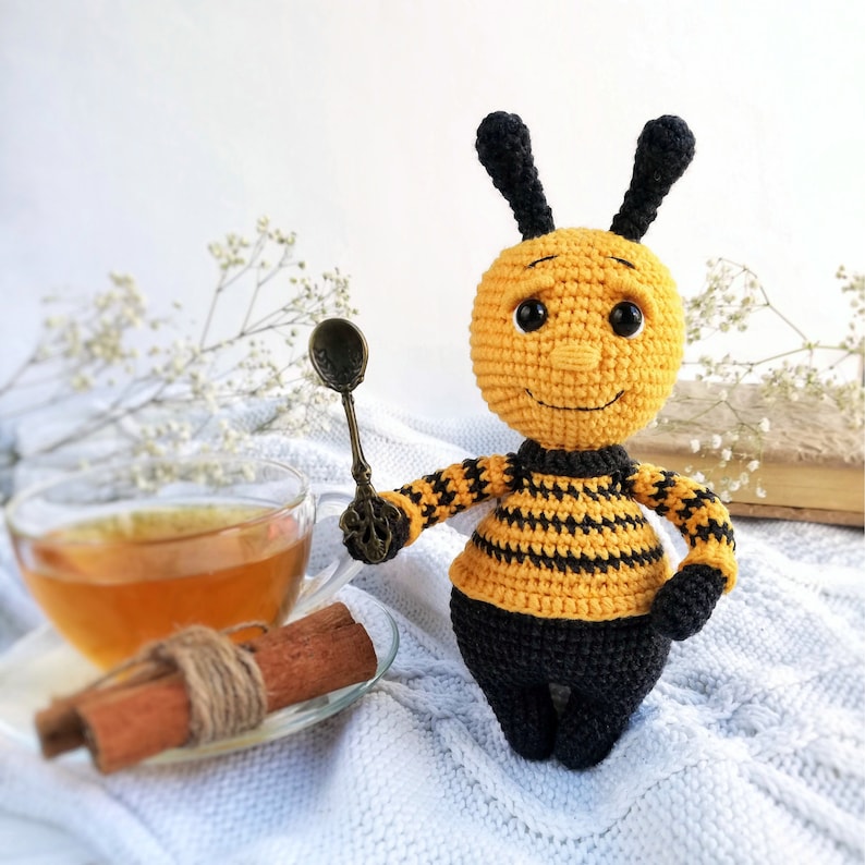 May include: A crocheted bee toy with black and yellow stripes, holding a small spoon. The bee has a smiling face with black eyes and a black nose. The bee is sitting on a white surface with a cup of tea and cinnamon sticks.