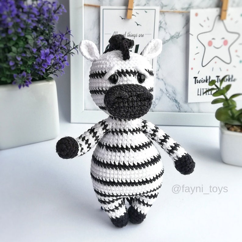 May include: A crocheted zebra toy with black and white stripes. The zebra has a black nose and black eyes. The toy is standing on a white surface.