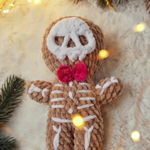 May include: A handmade, plush skeleton doll with a brown body and white bone details. The doll has a white skull face and a bright pink bow tie. The soft toy is designed to look like a gingerbread man.