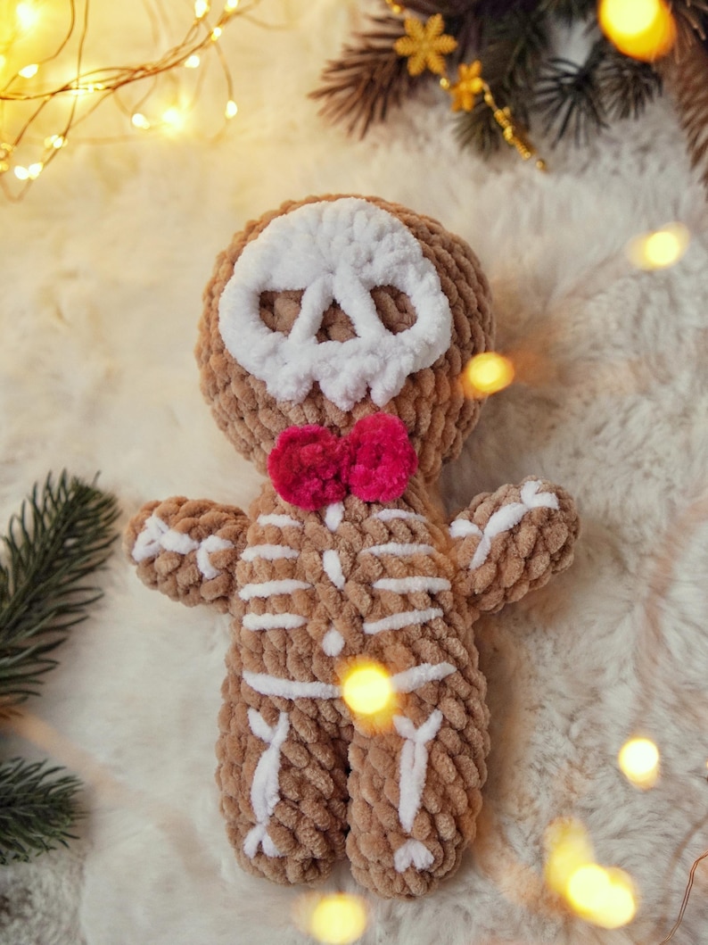 May include: A handmade, plush skeleton doll with a brown body and white bone details. The doll has a white skull face and a bright pink bow tie. The soft toy is designed to look like a gingerbread man.
