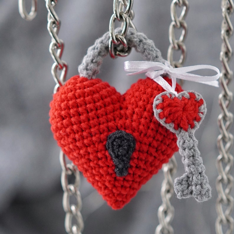 May include: A crocheted red heart-shaped padlock with a gray crocheted key attached. The padlock is hanging from a silver chain.