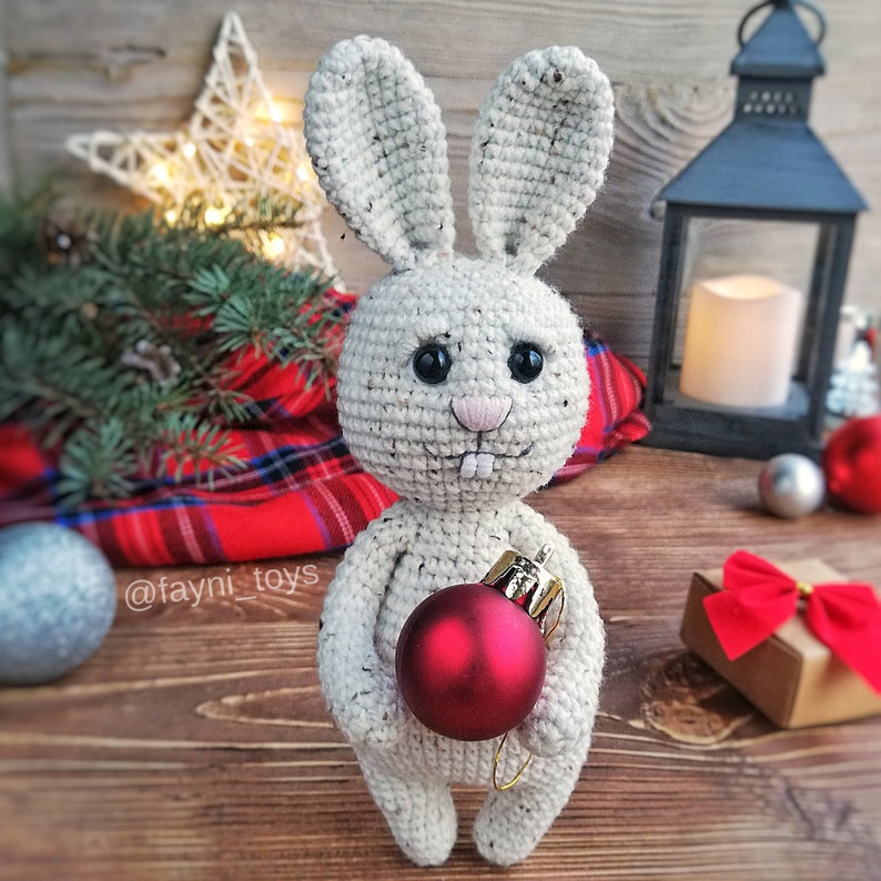 May include: A crocheted white bunny rabbit holding a red Christmas ornament. The bunny has black eyes and a pink nose. The bunny is standing on a wooden surface with a plaid blanket and a lantern in the background. @fayni_toys