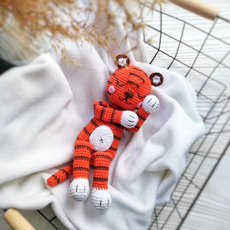 May include: A crocheted orange and white tiger stuffed animal with closed eyes, lying on a white blanket.