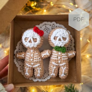 May include: Two crocheted skeleton dolls in a cardboard box. The dolls are tan with white skeleton detailing, white skull faces, and small accessories. One doll has a red bow, and the other has a green scarf. The box sits on a white doily.