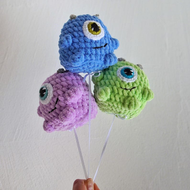 May include: Three crocheted plush monster balloons in blue, purple, and green with one large eye each. The balloons are tied together with white ribbon.