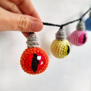 May include: A string of crocheted lightbulb ornaments with different colored eyes. The lightbulbs are connected with black string. The lightbulbs are red, yellow, pink, and blue.