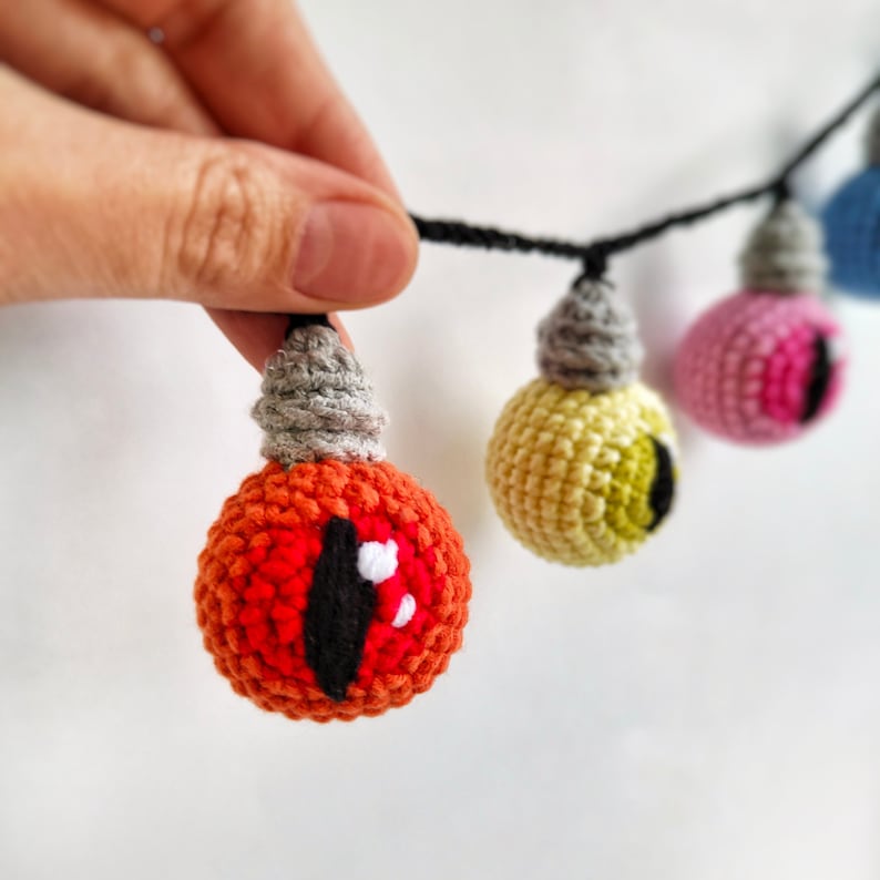 May include: A string of crocheted lightbulb ornaments with different colored eyes. The lightbulbs are connected with black string. The lightbulbs are red, yellow, pink, and blue.