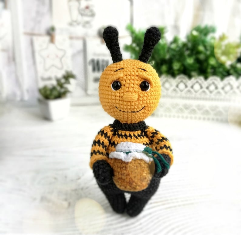 May include: A crocheted bee plush toy holding a pot of honey. The bee is yellow and black with a smiling face and black eyes. The honey pot is yellow and brown.