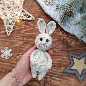 May include: A white crocheted bunny rabbit toy with black eyes and a pink nose. The bunny is being held in a person's hand.