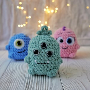 May include: Three crocheted plush toys in the shape of monsters. The first monster is blue with one large eye. The second monster is green with three eyes. The third monster is pink with two large eyes and a smiling mouth.