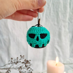 May include: A crocheted green apple with a black skull face. The apple is hanging from a string.