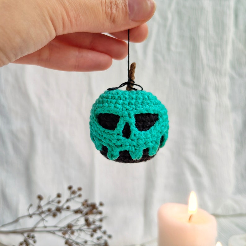 May include: A crocheted green apple with a black skull face. The apple is hanging from a string.