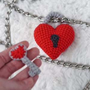 May include: A red crocheted heart-shaped padlock with a gray keyhole and a matching gray crocheted key. The padlock is attached to a silver chain.