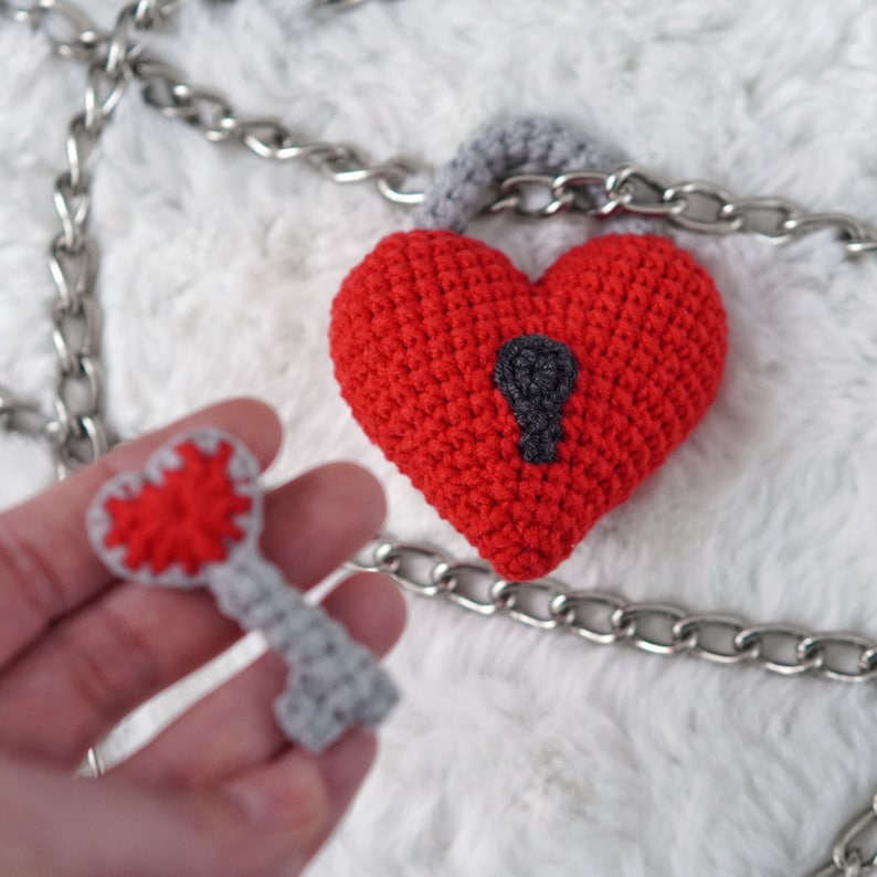 May include: A red crocheted heart-shaped padlock with a gray keyhole and a matching gray crocheted key. The padlock is attached to a silver chain.