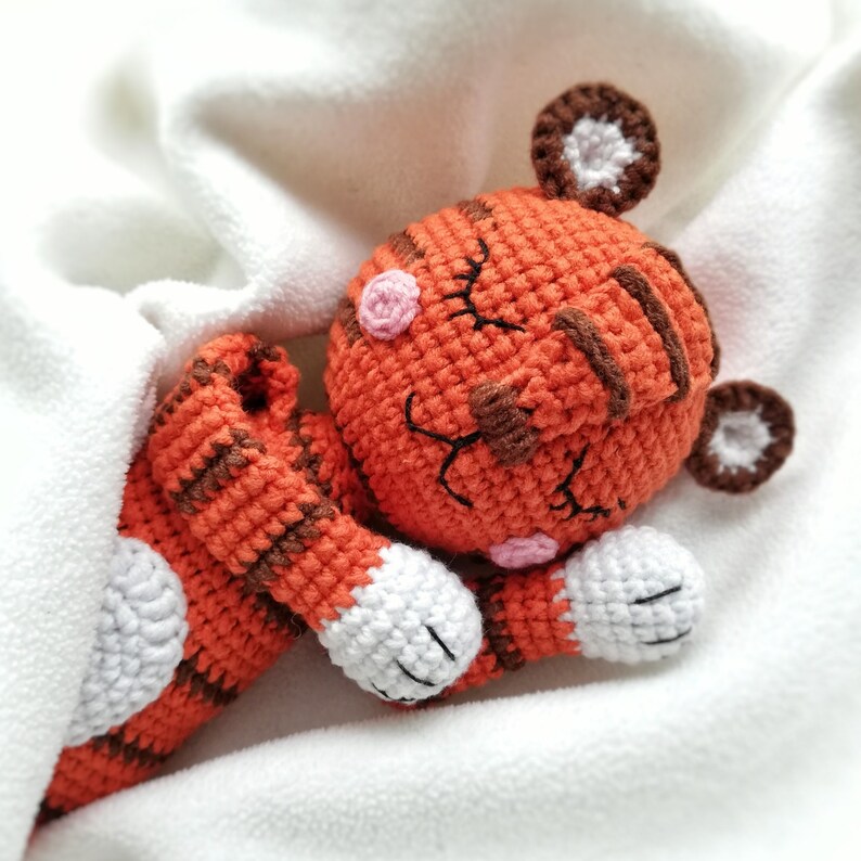May include: A crocheted orange and brown tiger stuffed animal with white accents is sleeping on a white blanket. The tiger has closed eyes and a pink nose.