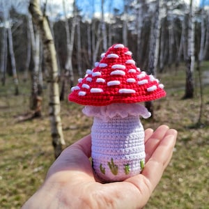 May include: A handmade crochet mushroom with a red cap and white spots. The stem is white with green embroidered grass details. The mushroom is held in a hand against a blurred forest background.