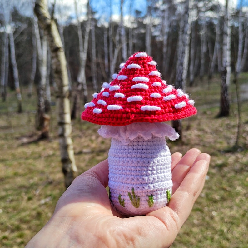 May include: A handmade crochet mushroom with a red cap and white spots. The stem is white with green embroidered grass details. The mushroom is held in a hand against a blurred forest background.
