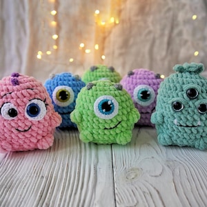 May include: Six crocheted plush toys shaped like monsters with different colors and facial features. The monsters are pink, blue, green, purple, and teal.