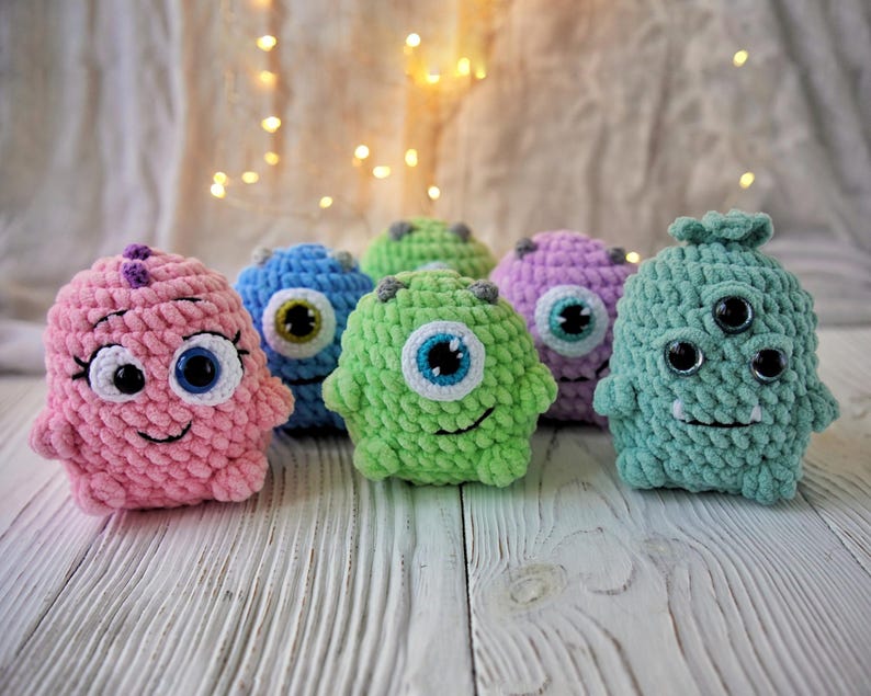 May include: Six crocheted plush toys shaped like monsters with different colors and facial features. The monsters are pink, blue, green, purple, and teal.
