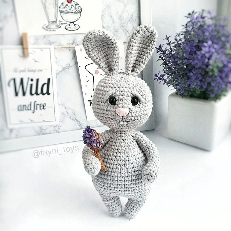 May include: A gray crocheted bunny rabbit holding a small bouquet of purple flowers. The bunny has black eyes and a pink nose.