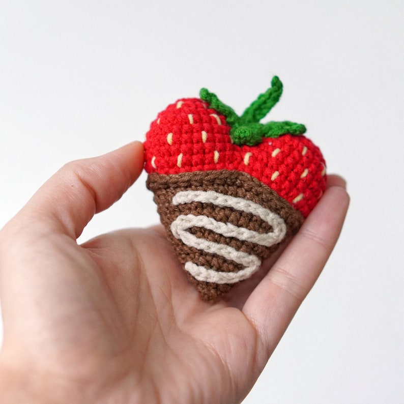 May include: A crocheted strawberry with a chocolate swirl design. The strawberry is red with green leaves and the chocolate is brown with white swirls.