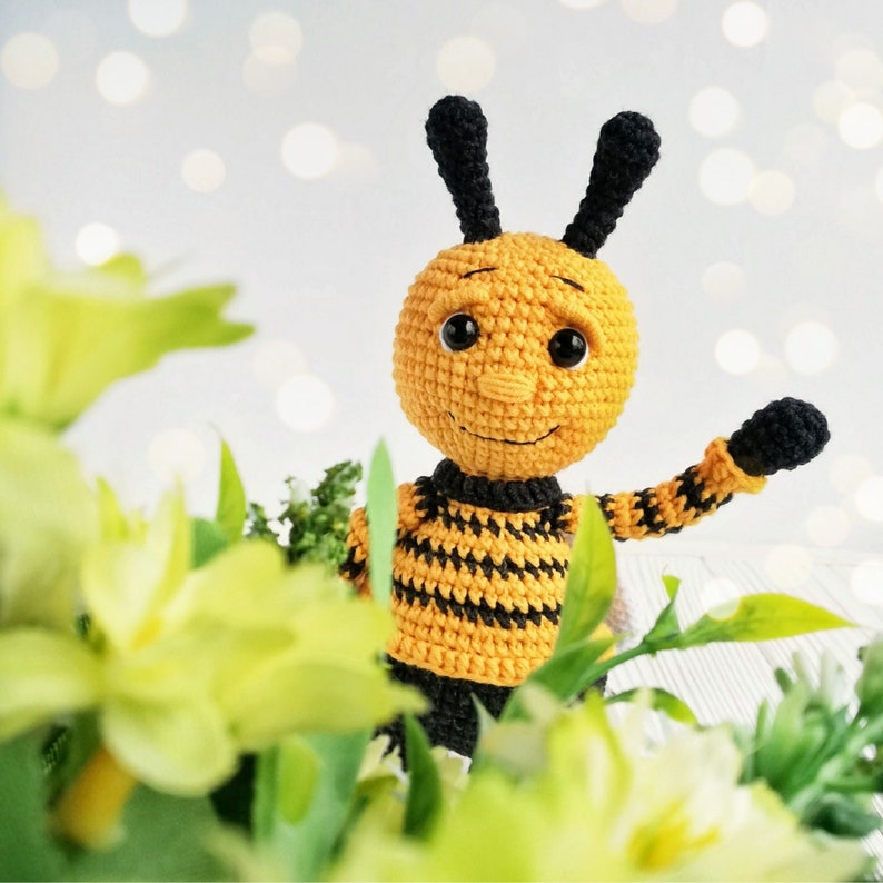 May include: A crocheted bee toy with black and yellow stripes, black eyes, and a smiling face. The bee is holding its right arm up in a waving motion.