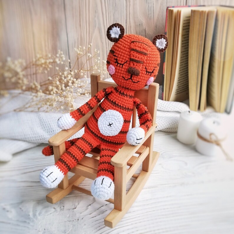 May include: A crocheted orange and white tiger plush toy with closed eyes sits in a wooden rocking chair. The chair is on a white surface.