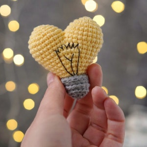 May include: A crocheted lightbulb shaped like a heart in yellow with a gray base. The lightbulb is held in a hand.
