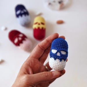 May include: A blue and white crocheted pill shaped toy with a skull design. The toy is being held in a hand.