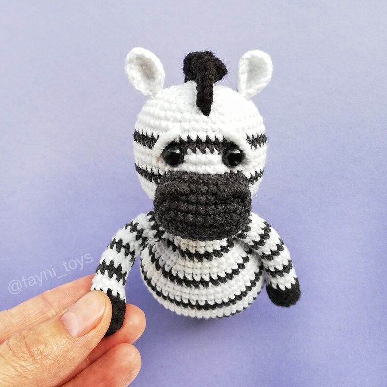 May include: A crocheted zebra toy with black and white stripes. The zebra has black eyes and a black nose. The toy is sitting on a purple surface.