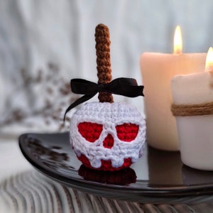 May include: A crocheted apple with a white skull and red eyes. The apple is on a black plate and there are two white candles in the background.