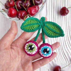 Puede incluir: Dos cerezas de ganchillo con hojas verdes. Las cerezas son rojas y tienen un diseño de ojo blanco y azul.