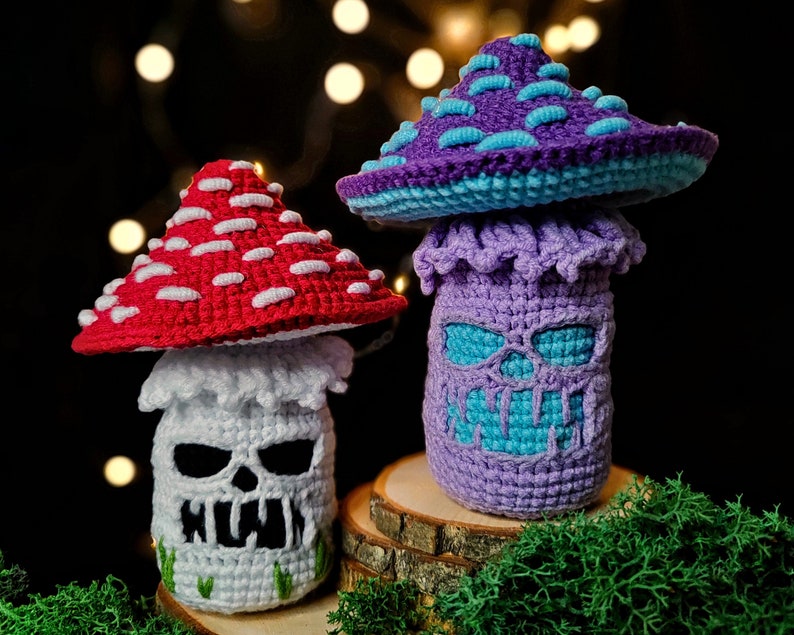 May include: Two crocheted mushroom figures. One has a red cap with white spots and a white stem with a skull face. The other has a purple cap with blue spots and a purple stem with a teal skull face. The mushrooms are on a wooden base.
