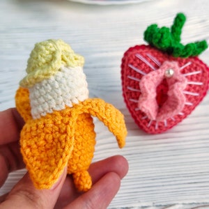 Op de afbeelding: Een gehaakte banaan met een gele bovenkant en een oranje lichaam, en een gehaakte aardbei met een roze hart en groene bladeren.