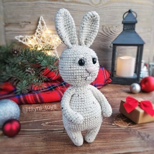 May include: A white crocheted bunny rabbit toy with black eyes and a pink nose. The bunny is standing on a wooden surface with a red and black plaid blanket and a Christmas-themed background.