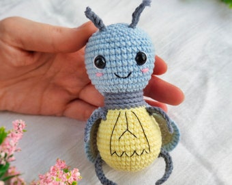 May include: A crocheted firefly toy with a light blue head, yellow body, and black legs. The toy has a smiling face with black eyes and pink cheeks.