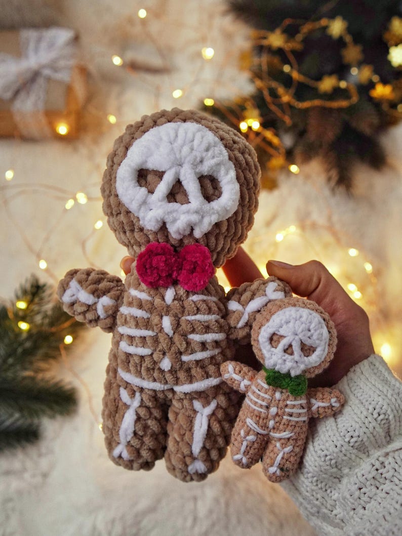 May include: Two crocheted skeleton dolls. The larger doll is brown with white bone detailing, a white skull face, and a pink bow tie. The smaller doll is tan with white bone detailing, a white skull face, and a green bow tie.
