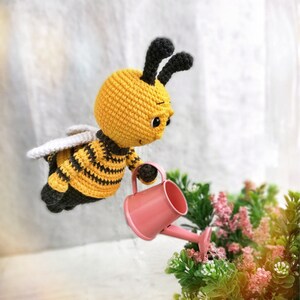 May include: A crocheted bee toy holding a pink watering can. The bee is yellow and black with white wings. The bee is watering a small green plant with pink flowers.