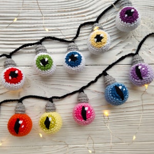 May include: A string of crocheted light bulbs shaped like eyeballs in various colors, including red, green, blue, yellow, pink, and purple. The bulbs are strung together with black cord and are decorated with black pupils. The string of lights is hanging on a white wooden surface.